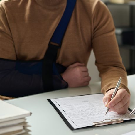 Nahaufnahme eines Mannes, der einen Arm in einer Schlinge befestigt hat und mit der anderen ein Formular für medizinische Versicherung ausfüllt