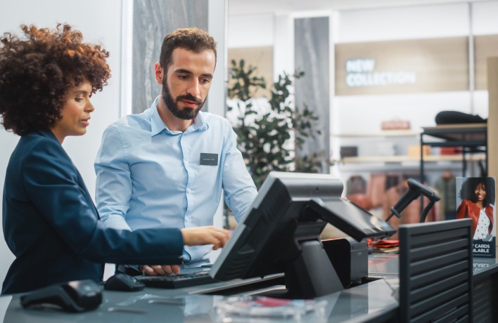 Kasse eines Einkaufsgeschäfts mit zwei Mitarbeitenden: Vordere Person zeigt auf den Bildschirm